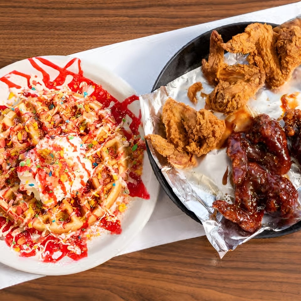 Waffles with whipped cream, sprinkles, and strawberry syrup next to fried chicken and BBQ wings.