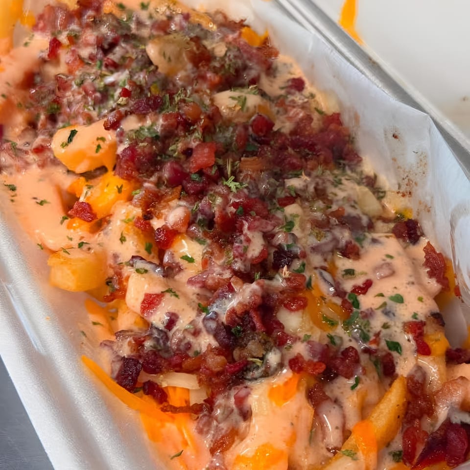 Close-up of loaded fries with melted cheese, bacon bits, and creamy sauce, garnished with herbs.